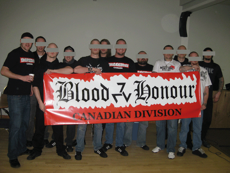 12 people with their faces sensored hold a banner saying "Blood & Honour CANADIAN DIVISION". The ampersand is replaced by a Triskeles styled to resemble three sevens.