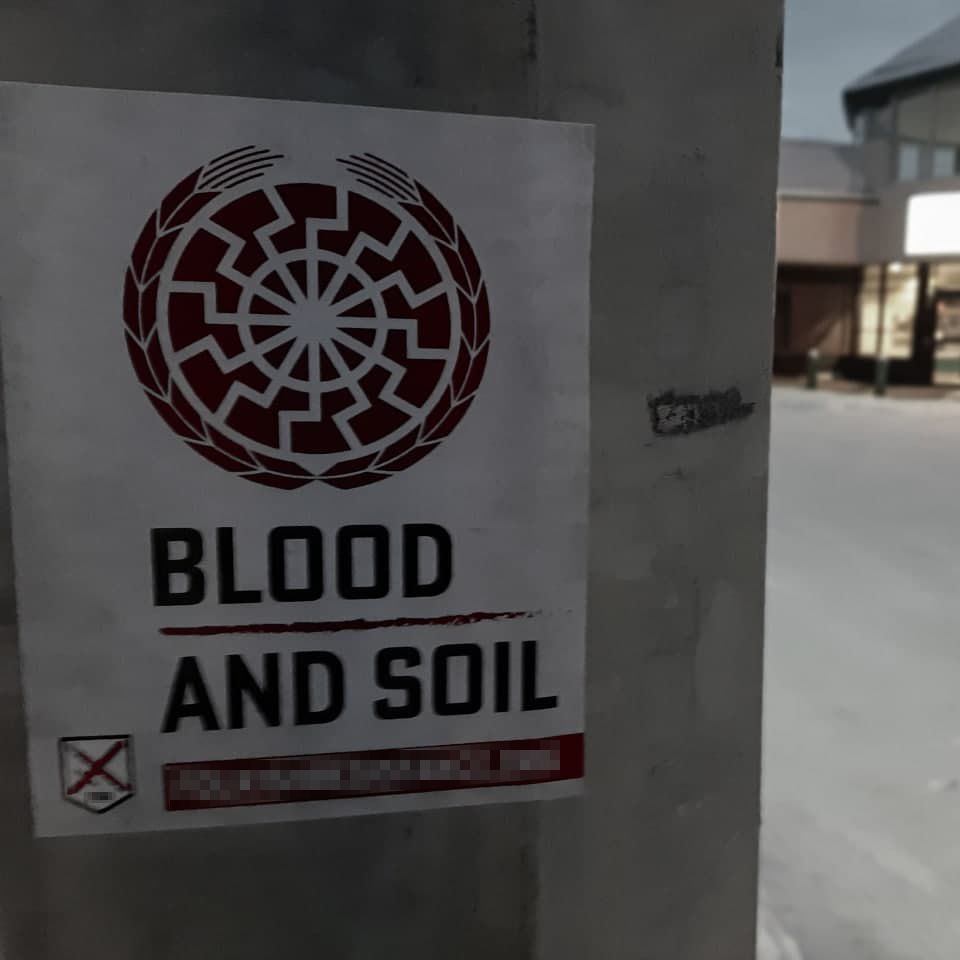 Folkish Resistance Movement's "Blood and Soil" poster on a steel structure. A single-floor building with a large foyer appears in the background. A Black Sun (Sonnenrad) is placed in a wreath. Below is the text "BLOOD", a red line, and then "AND SOIL".