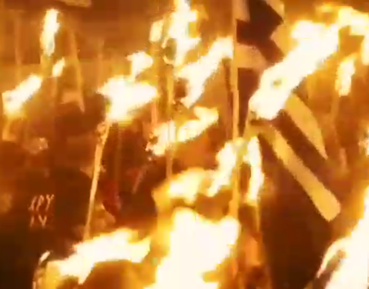 Image fixe d'une torche tiki marchant de près. Les hommes défilent avec des torches tiki hautes et un drapeau est levé. Les flammes occupent une grande partie du cadre.