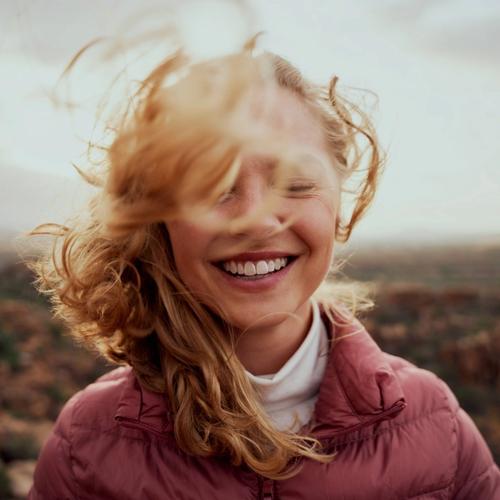En tjej står ute i ett öppet landskap. Håret blåser åt alla håll och hon ler stort. bild.