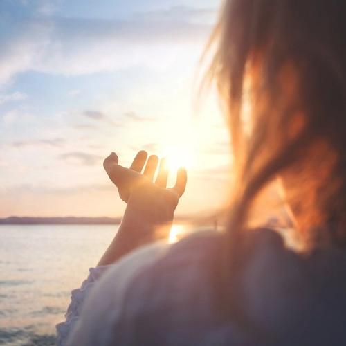 En kvinna står vid vattnet och håller ut handen så det ser ut som fingrarna rör vid solen. foto.