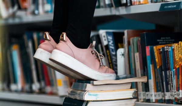 Noen med rosa skor står på bøker for å strekke seg enda høyere etter en bok i hyllen. bilde.