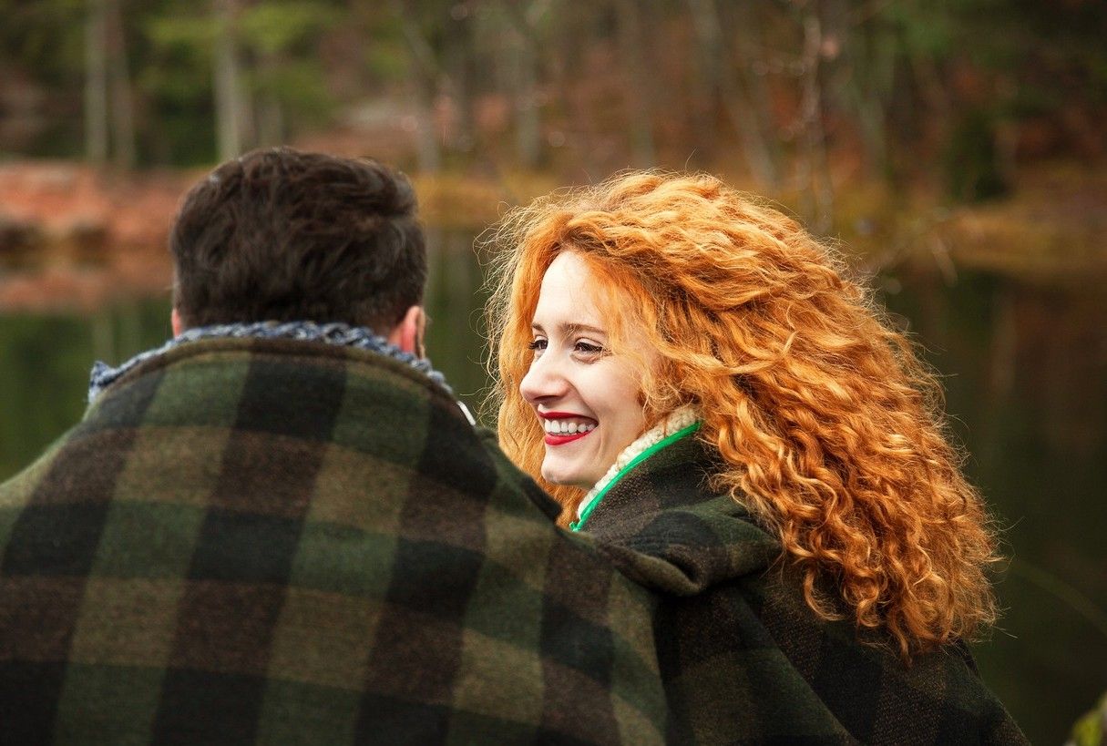 En man och en kvinna sitter ute med en stor filt om sig. Kvinnan har stort, lockigt, rött hår och hon ler stort mot mannen. foto.