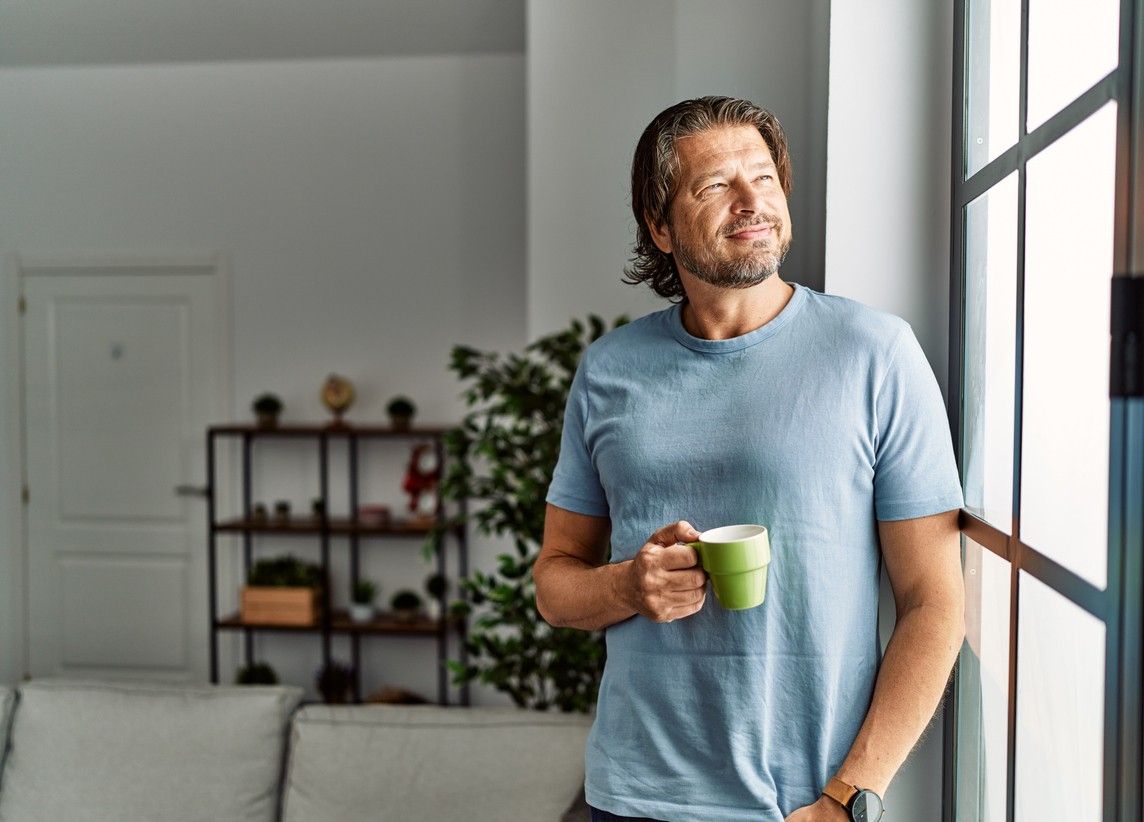 Man står med en kaffekopp i handen och ser ut genom fönstret. foto.