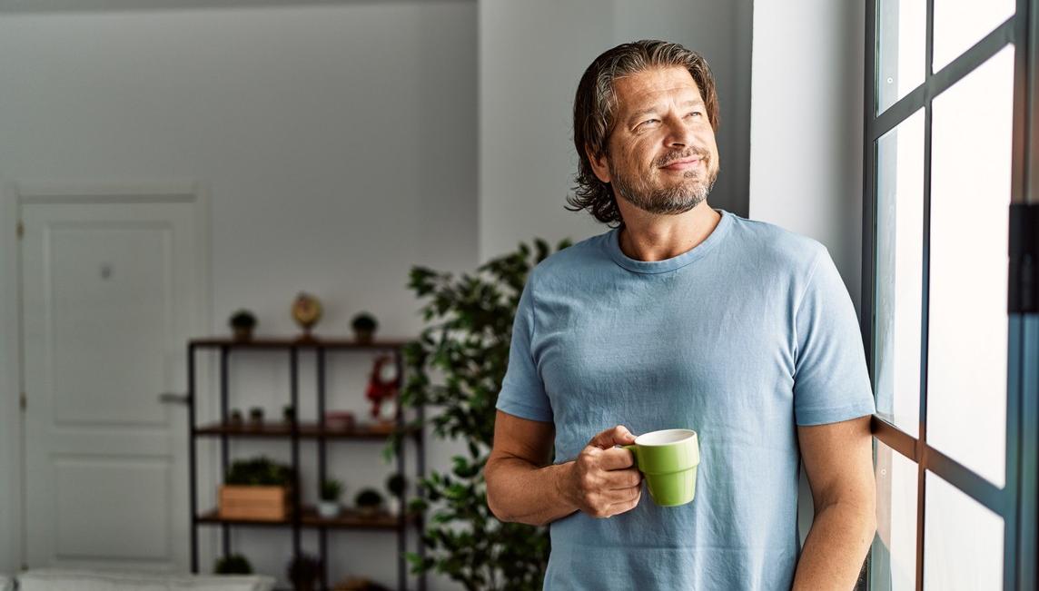 Man står med en kaffekopp i handen och ser ut genom fönstret. foto.