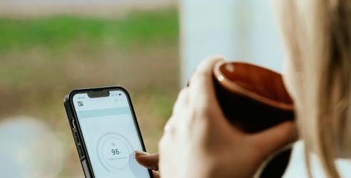 Kvinna står vid fönstret och ser på mobilen medan hon dricker kaffe