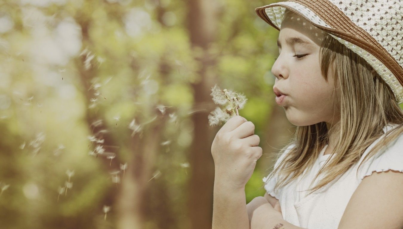 En jente i solhatt blåser på en løvetann. bilde.