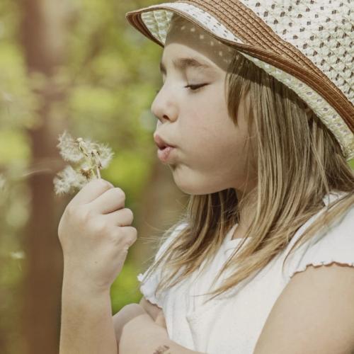 En jente i solhatt blåser på en løvetann. bilde.