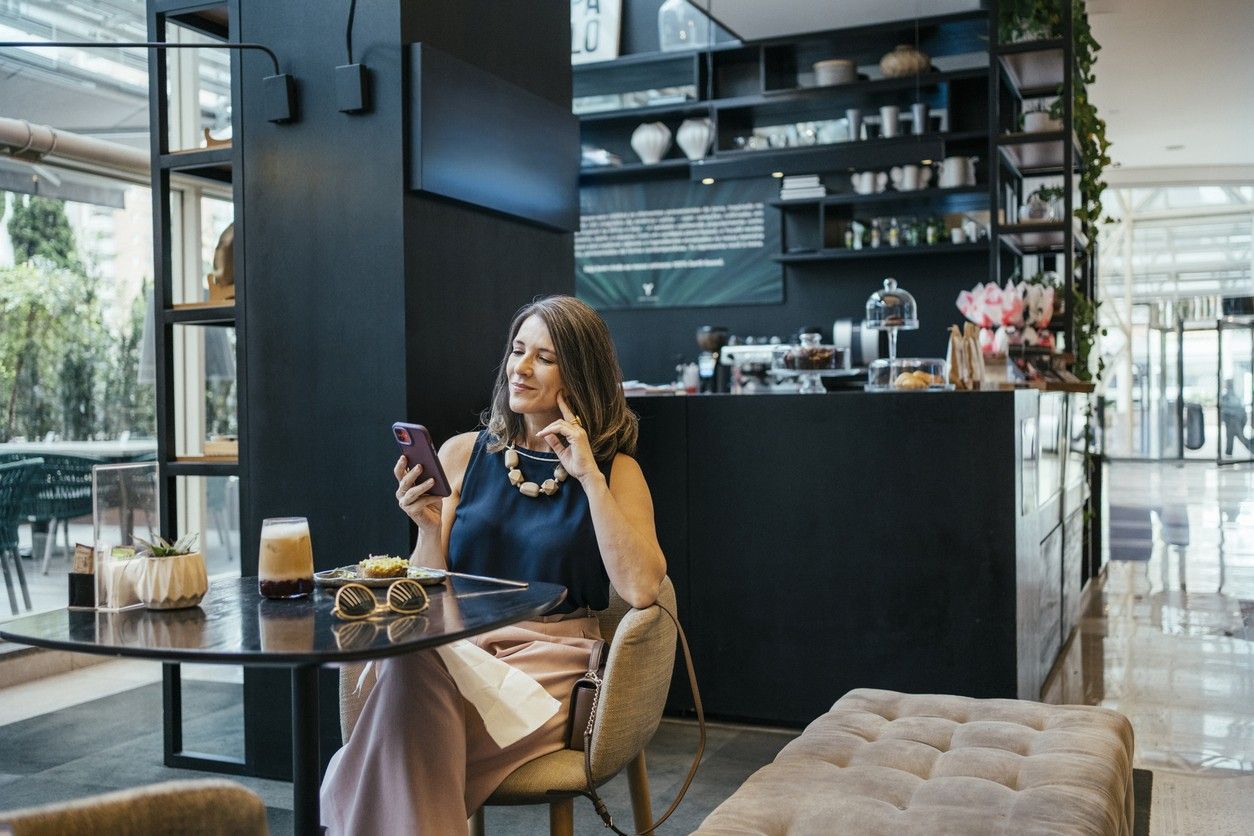Nainen istuu kahvilassa ja katsoo matkapuhelintaan. Valokuva.