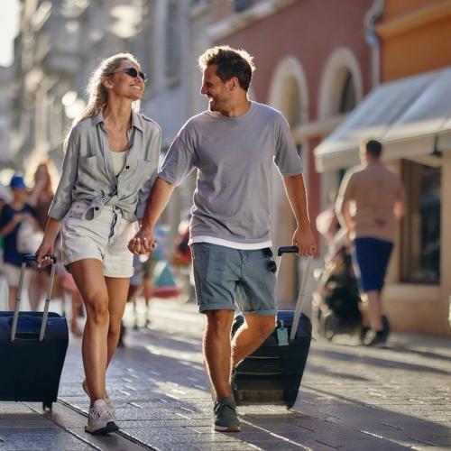 Ett par på resa drar varsin koffert bakom sig. De ler mot varandra. bild.