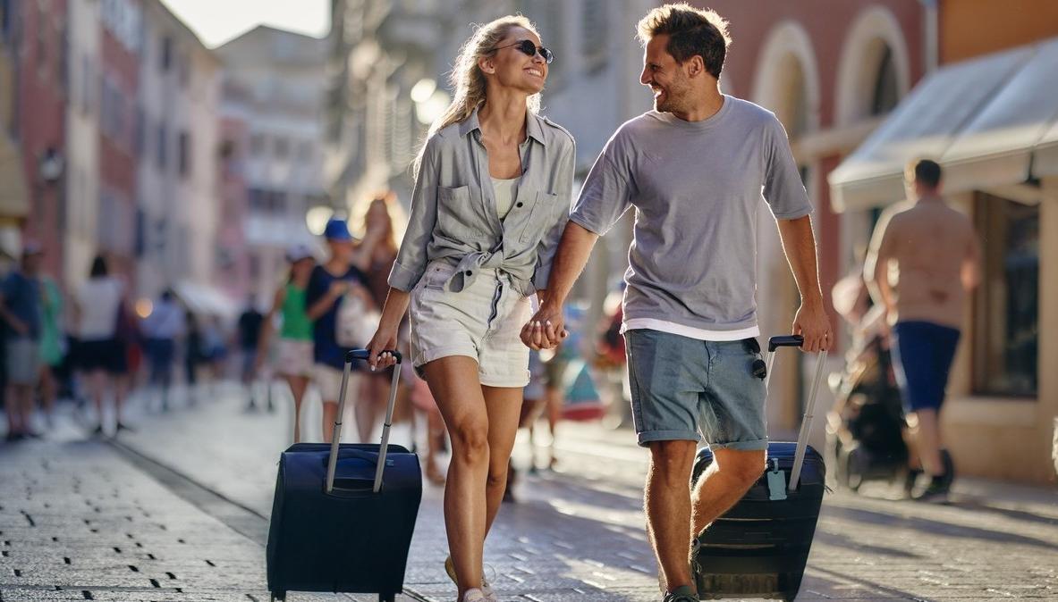 Ett par på resa drar varsin koffert bakom sig. De ler mot varandra. bild.