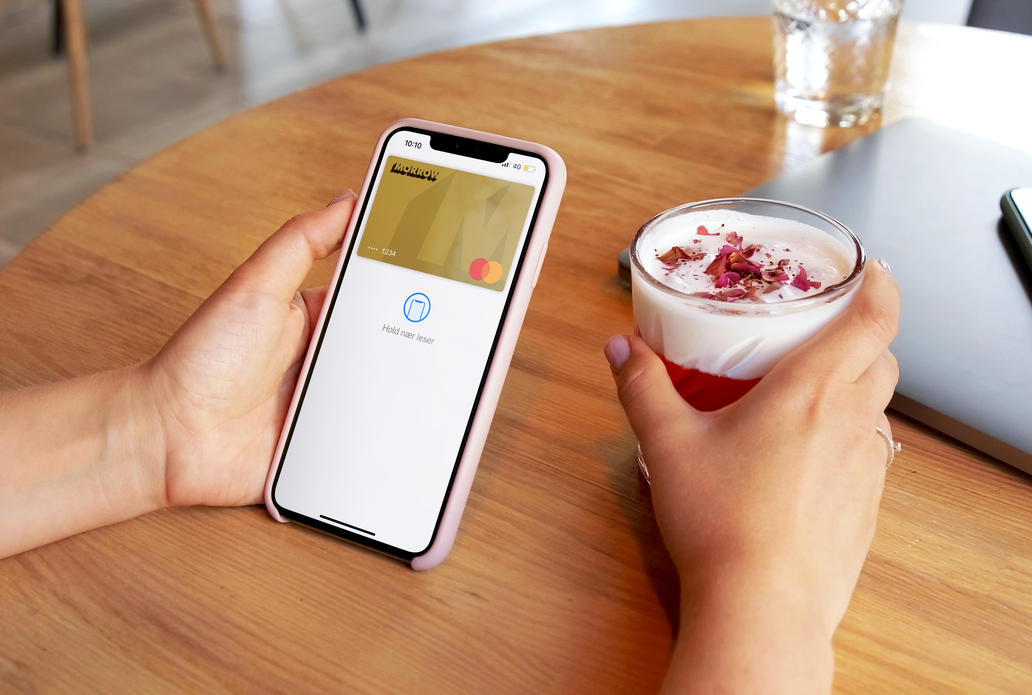 En hand håller mobil med Morrow guldkort i Apple Pay. Den andra handen håller en dryck med skummad mjölk och röd topping. foto.