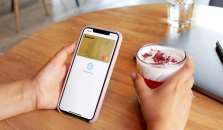 En hand håller mobil med Morrow guldkort i Apple Pay. Den andra handen håller en dryck med skummad mjölk och röd topping. foto.