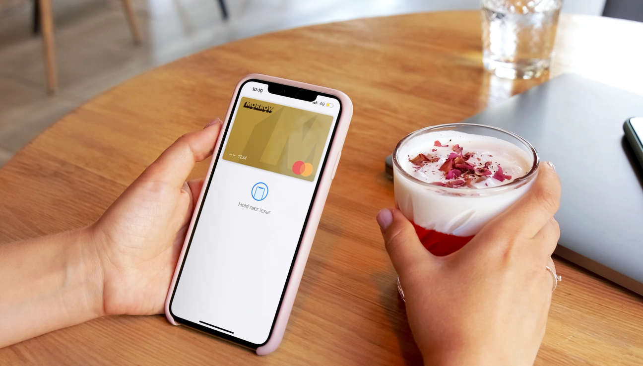 på cafe med Morrow Bank kredittkort i Apple Wallet i ena handen och en dryck i den andra. foto.