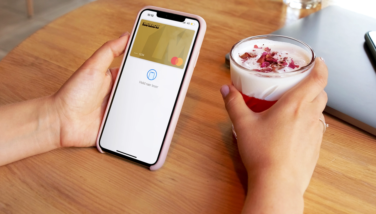 En hand håller mobil med Morrow guldkort i Apple Pay. Den andra handen håller en dryck med skummad mjölk och röd topping. foto.