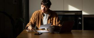 Man sitter ved et bord og jobber med pc, mobil og kaffekopp foran seg. Han har penn og papir.