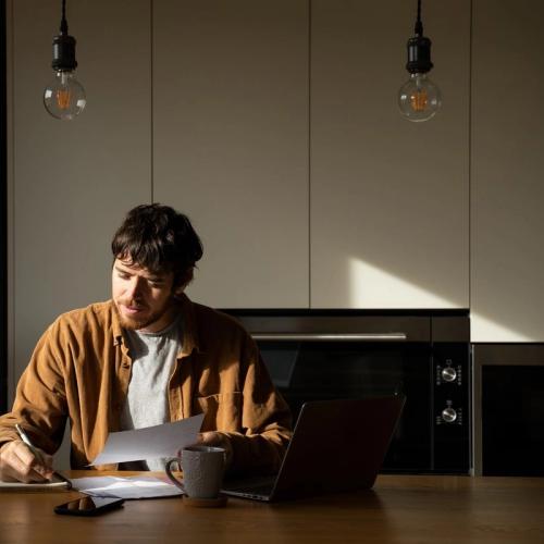 Man sitter ved et bord og jobber med pc, mobil og kaffekopp foran seg. Han har penn og papir.