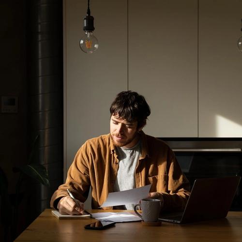 Man sitter ved et bord og jobber med pc, mobil og kaffekopp foran seg. Han har penn og papir.