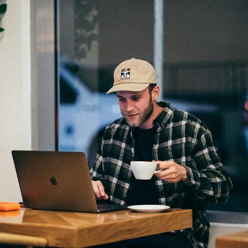En man sitter vid ett bord med en kaffekopp i handen och en mac framför sig