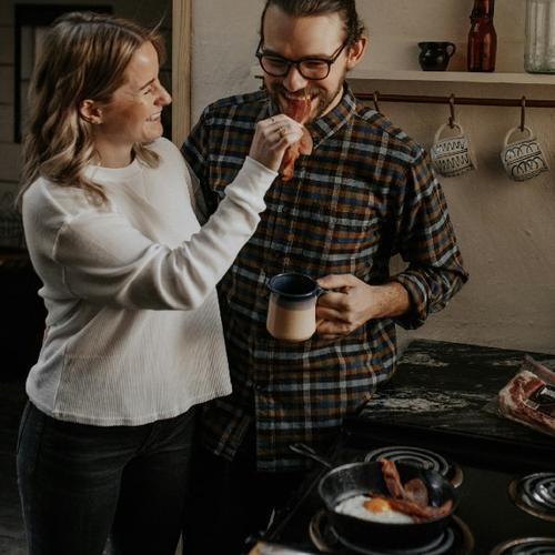 Ett par står vid spisen, hon matar honom med bacon. foto.