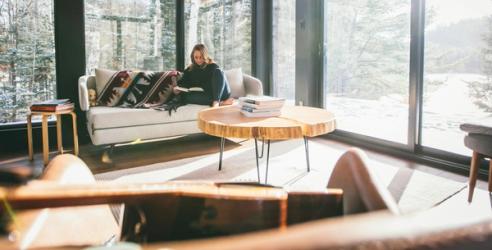 Stue med store vinduer. i sofaen sitter en dame og leser. bilde.