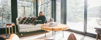 Stue med store vinduer. i sofaen sitter en dame og leser. bilde.