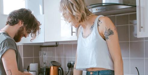 Et ungt par på kjøkkenet. Han står og skjærer noe. Hun sitter på kjøkkenbenken. bilde.
