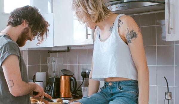 Et ungt par på kjøkkenet. Han står og skjærer noe. Hun sitter på kjøkkenbenken. bilde.