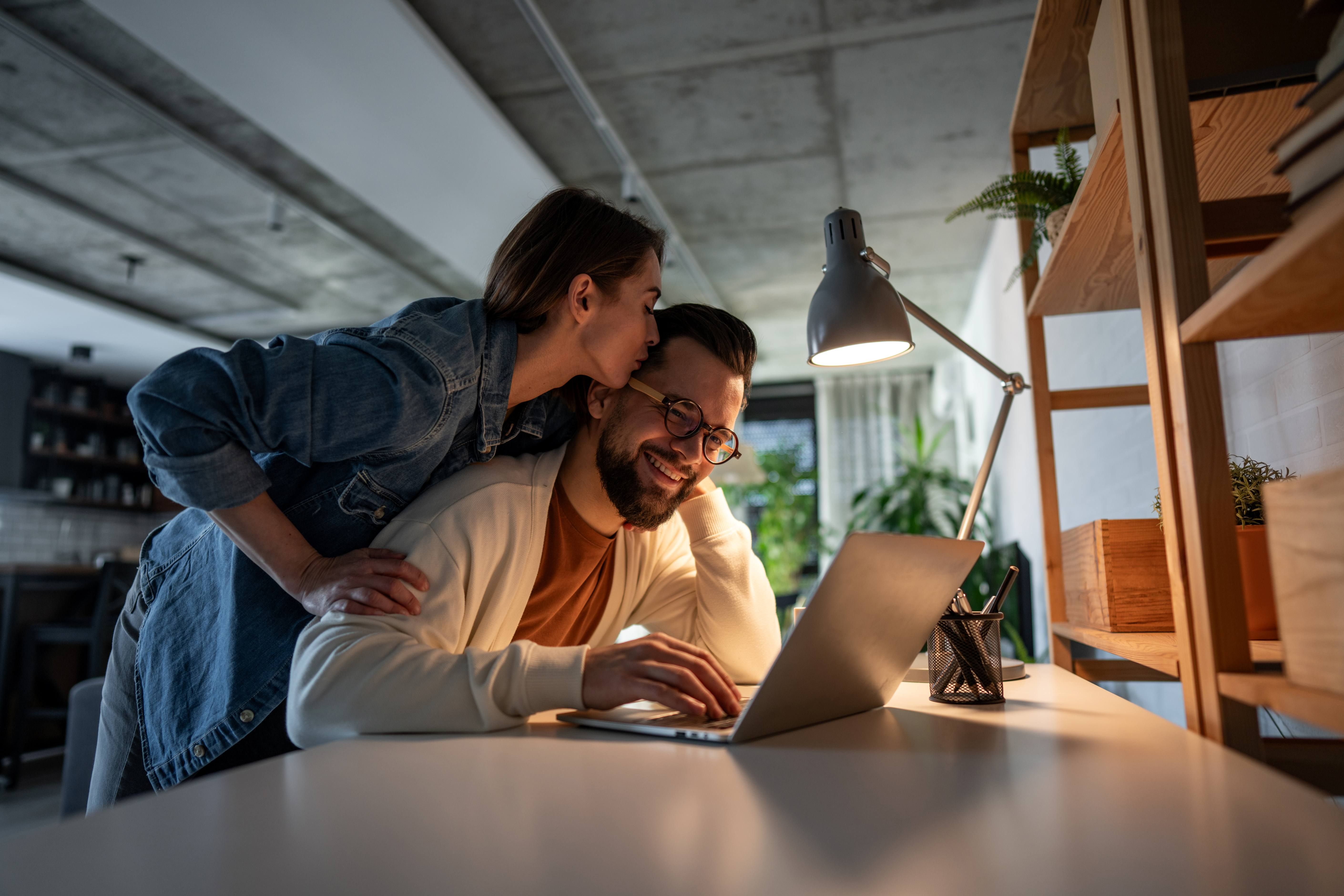 En man sitter med hånden på en Mac. Damen står bak ham og kysser ham på hodet. bilde.