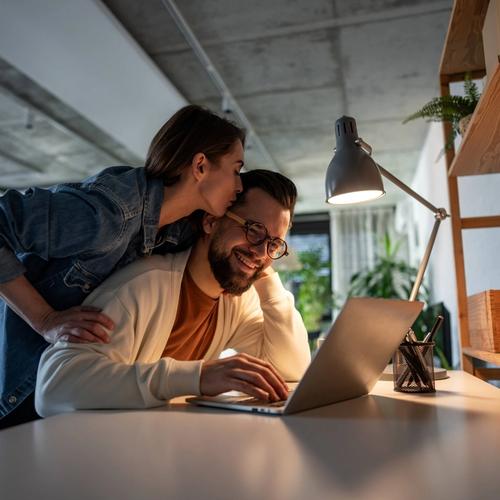 En man sitter med hånden på en Mac. Damen står bak ham og kysser ham på hodet. bilde.