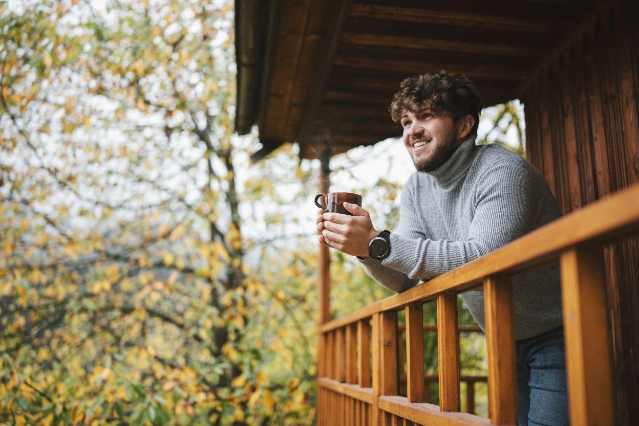 En man står ute på terrassen och lutar sig mot räcket med en kaffekopp i handen.