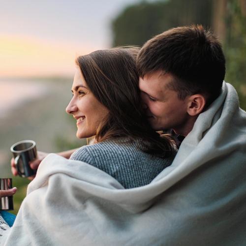 En man och en kvinna sitter ute. mannen har en filt om sig som han också lägger om kvinnans axlar. bild.