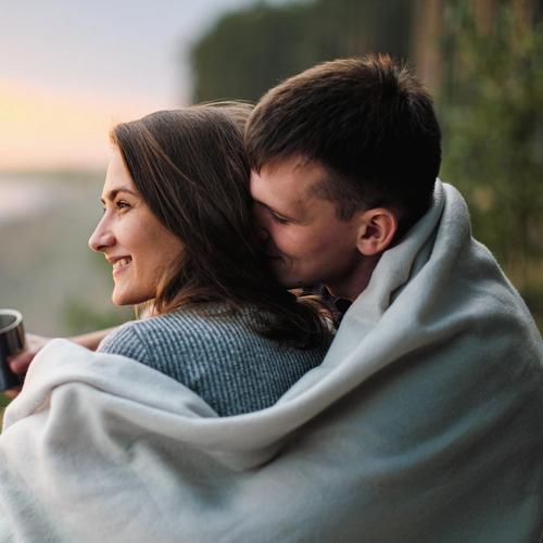 En man och en kvinna sitter ute. mannen har en filt om sig som han också lägger om kvinnans axlar. bild.