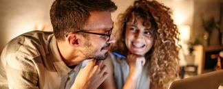 Mann og dame sitter ved pc. De ser på hverandre og smiler. bilde.