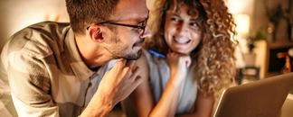 Mann og dame sitter ved pc. De ser på hverandre og smiler. bilde.