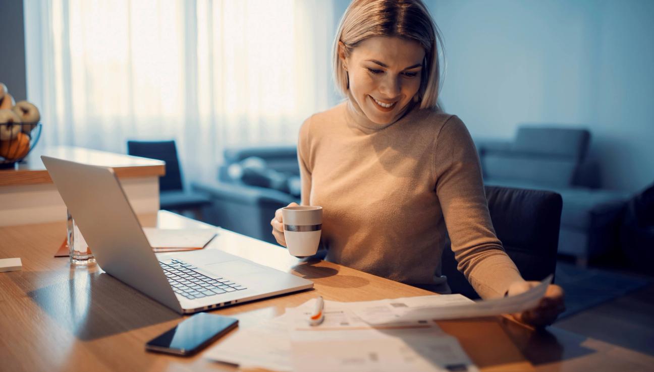 Dame sitter med kaffekopp og papirer med pc og mobil på bordet foran seg. bilde.