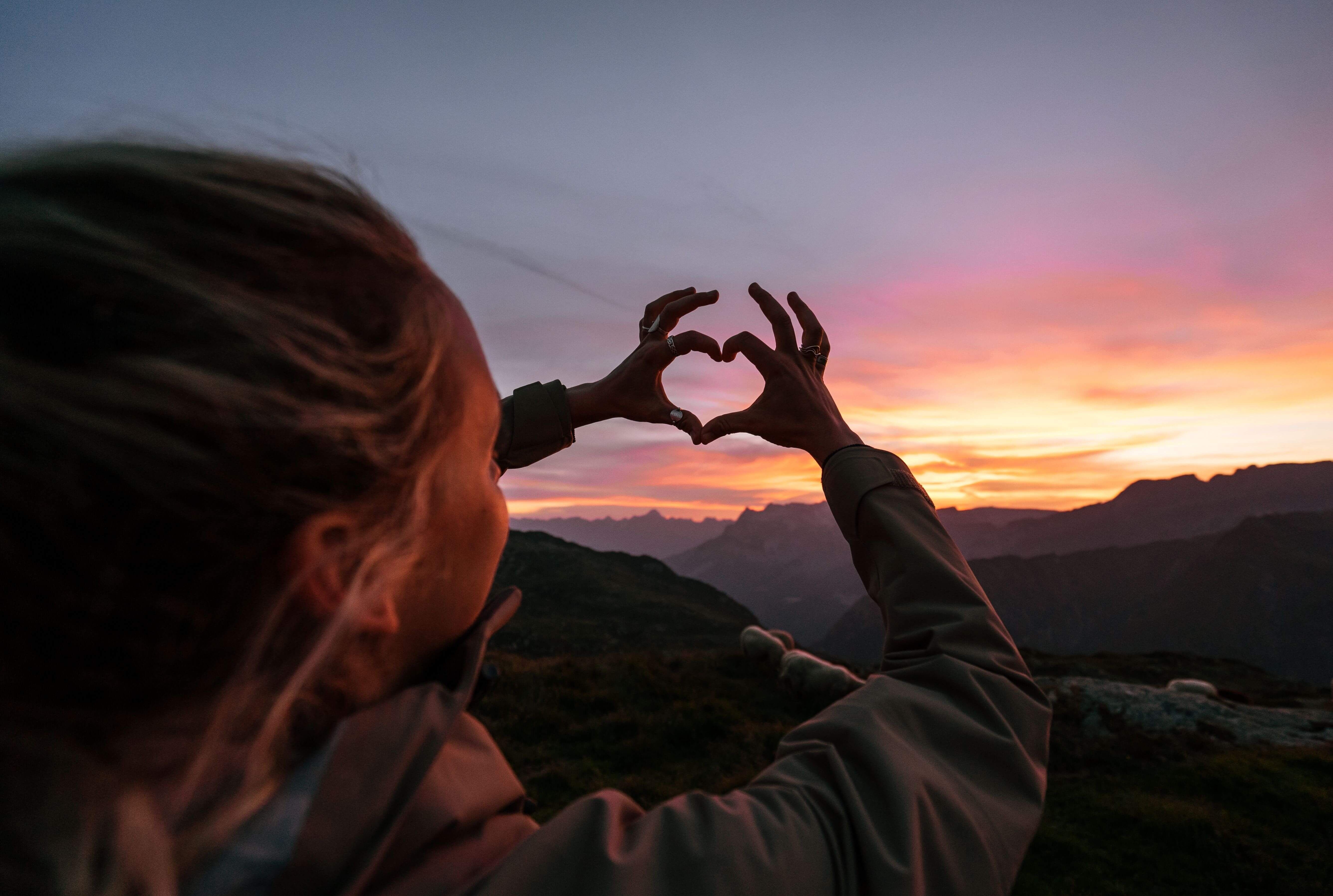 Kvinna håller händerna formade som ett hjärta mot solnedgången och den färgade himlen. foto.