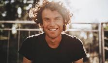 En man med lockigt hår ser mot kameran och ler. Solen skiner bakom honom. bild.