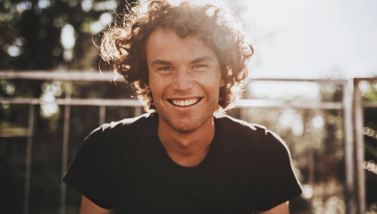 En man med lockigt hår ser mot kameran och ler. Solen skiner bakom honom. bild.