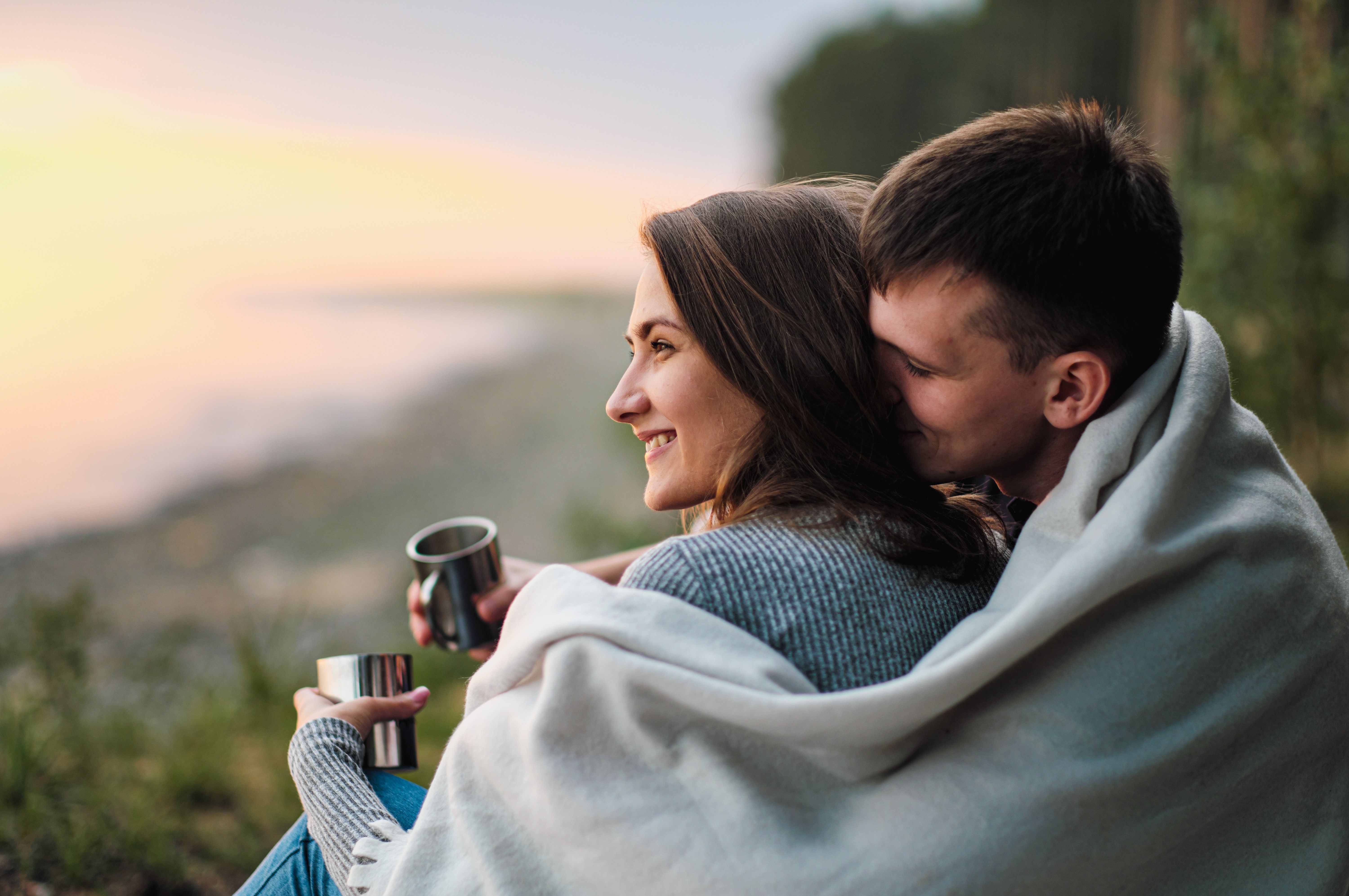 Mies istuu naisen takana, pitäen vilttiä heidän molempien ympärillä. Valokuva.