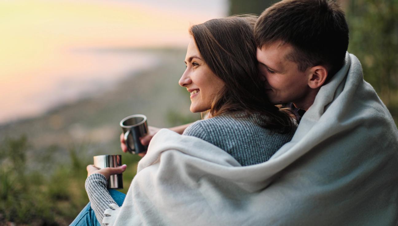 Mies istuu naisen takana, pitäen vilttiä heidän molempien ympärillä. Valokuva.