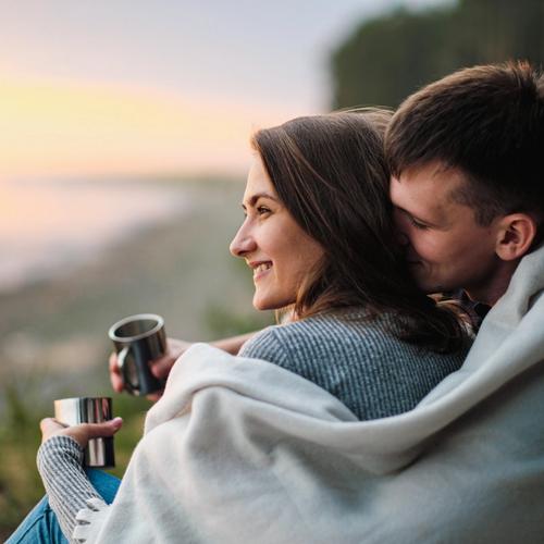 Mies istuu naisen takana, pitäen vilttiä heidän molempien ympärillä. Valokuva.