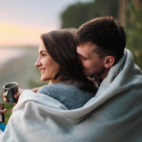 Mies istuu naisen takana, pitäen vilttiä heidän molempien ympärillä. Valokuva.