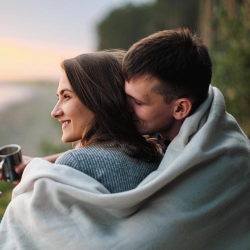 Mies istuu naisen takana, pitäen vilttiä heidän molempien ympärillä. Valokuva.