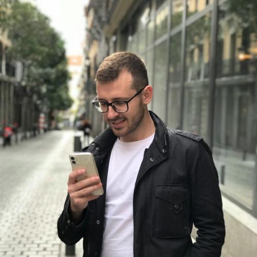 Mann med briller og svart jakke står på fortauet og leser på sin mobil. bilde.