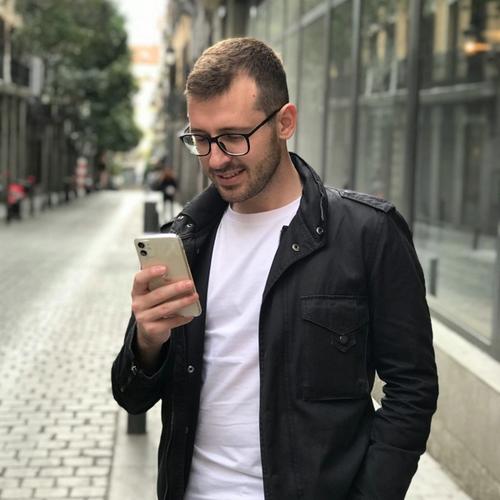 Mann med briller og svart jakke står på fortauet og leser på sin mobil. bilde.