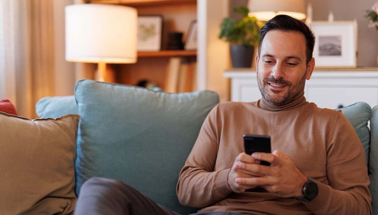 En man sitter i soffan och nätsurfar på telefonen. foto.