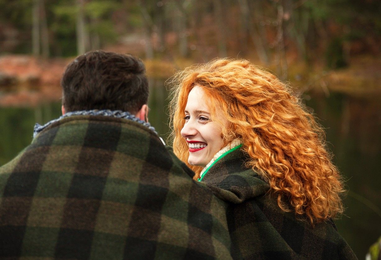 En mann og en dame sitter ute med et stort pledd om seg. Damen har stort krøllete rødt hår, og smiler mot mannen. bilde.
