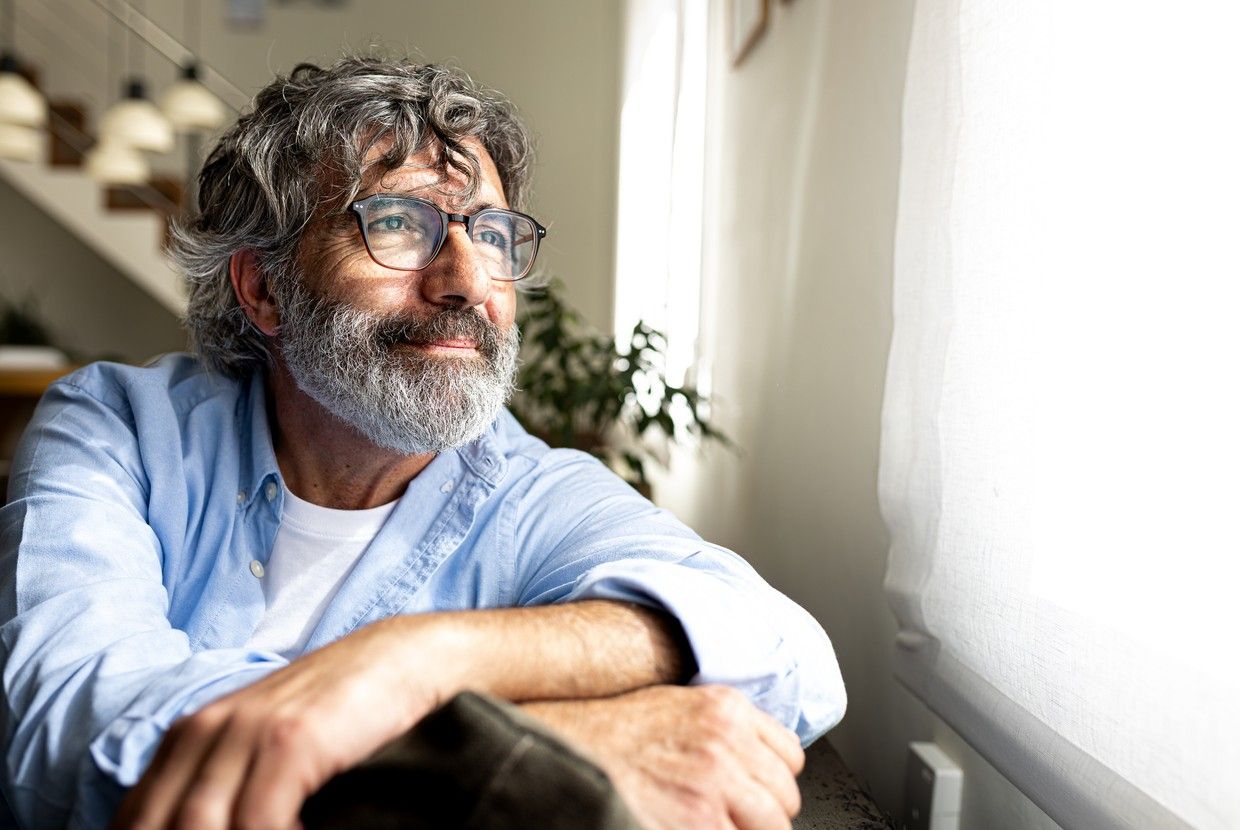 En mann sitter ved vinduet og ser ut. Solen skinner inn gjennom gardinen. bilde.