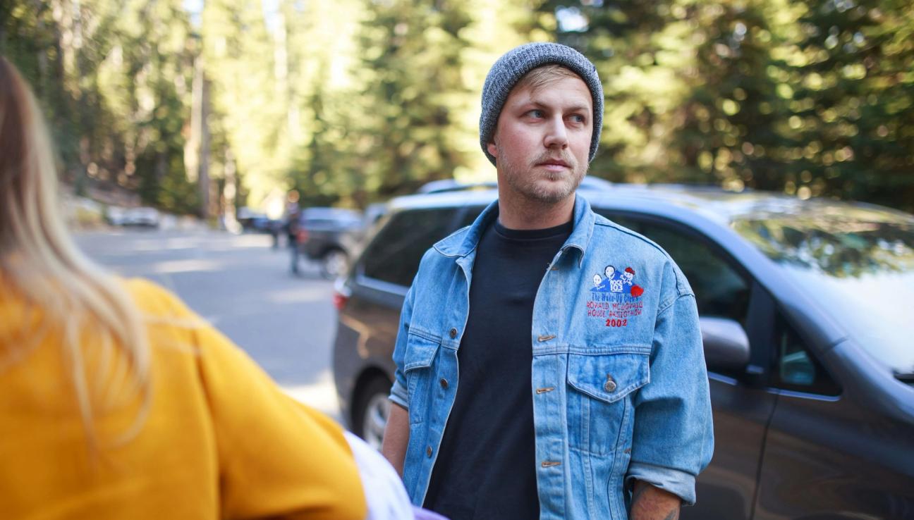 En man med jeansjacka står framför en bil och tittar bortåt